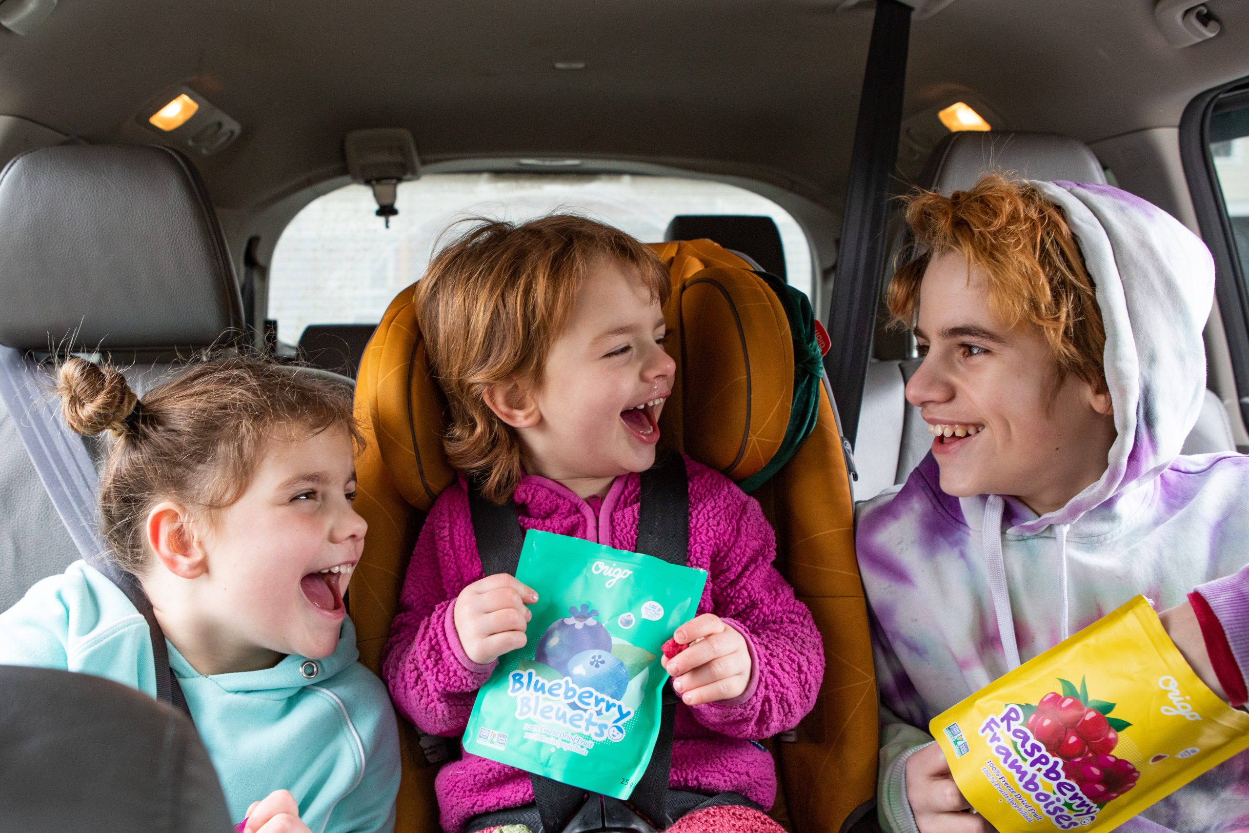 Three children laughing in the back seat while holding Origo blueberry and raspberry freeze-dried fruit pouches